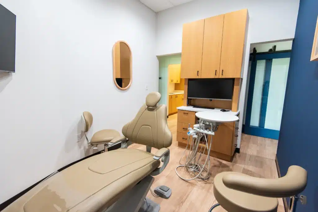 A modern dental exam room with beige dental chairs, a mounted screen, a round mirror, wood cabinets, and dental equipment on a tray. The room has light wood floors and a minimalist design.