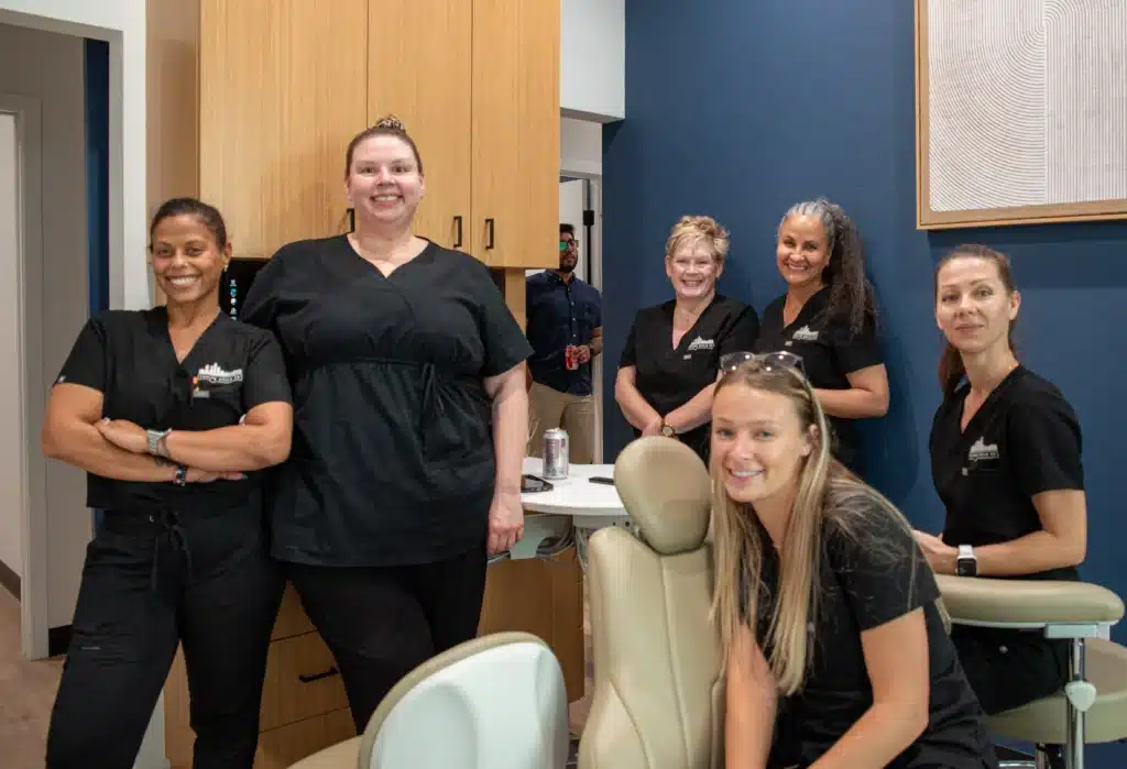 Six women in black scrubs smile and pose together in a modern dental office, with dental chairs and cabinets visible in the background.