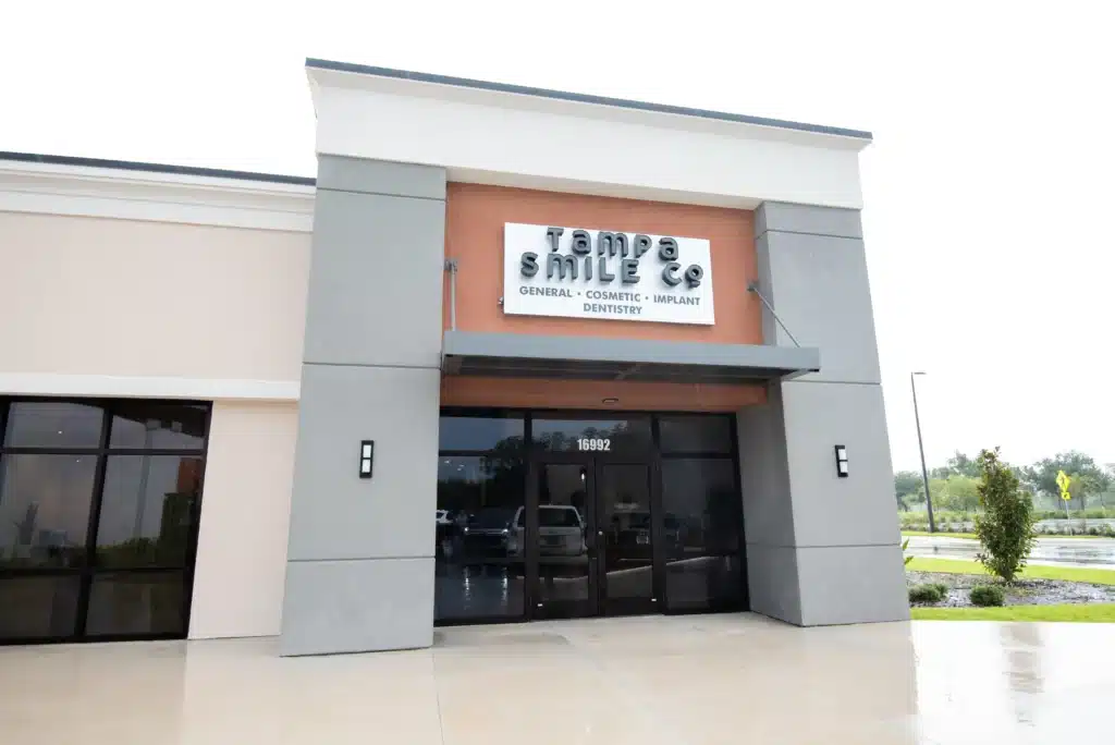 Front view of a modern dental office building with a sign reading Tampa Smiles: General, Cosmetic, Implant Dentistry above the entrance on a cloudy day.