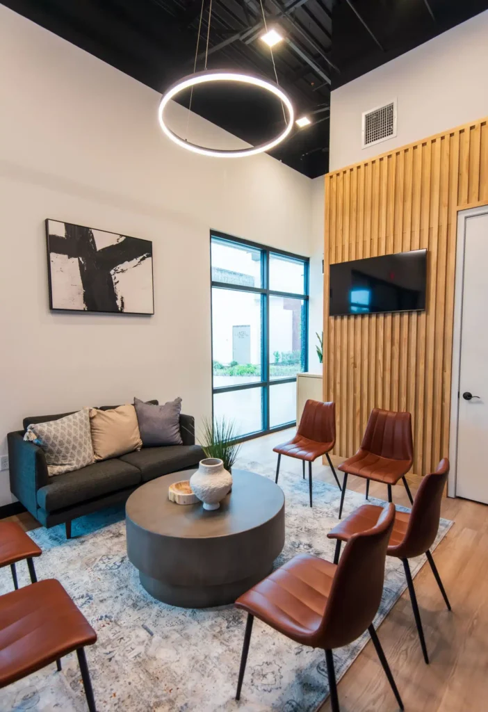 Modern lounge with a gray sofa, brown chairs around a round table, abstract wall art, potted plants, a large rug, circular ceiling light, wall-mounted TV, and wood slat accent wall. Large windows provide natural light.