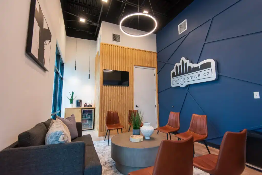 A modern waiting room with brown leather chairs, a gray sofa, a round coffee table, a blue accent wall with a sign reading Tampa Smile Co, wooden wall panels, and contemporary lighting.