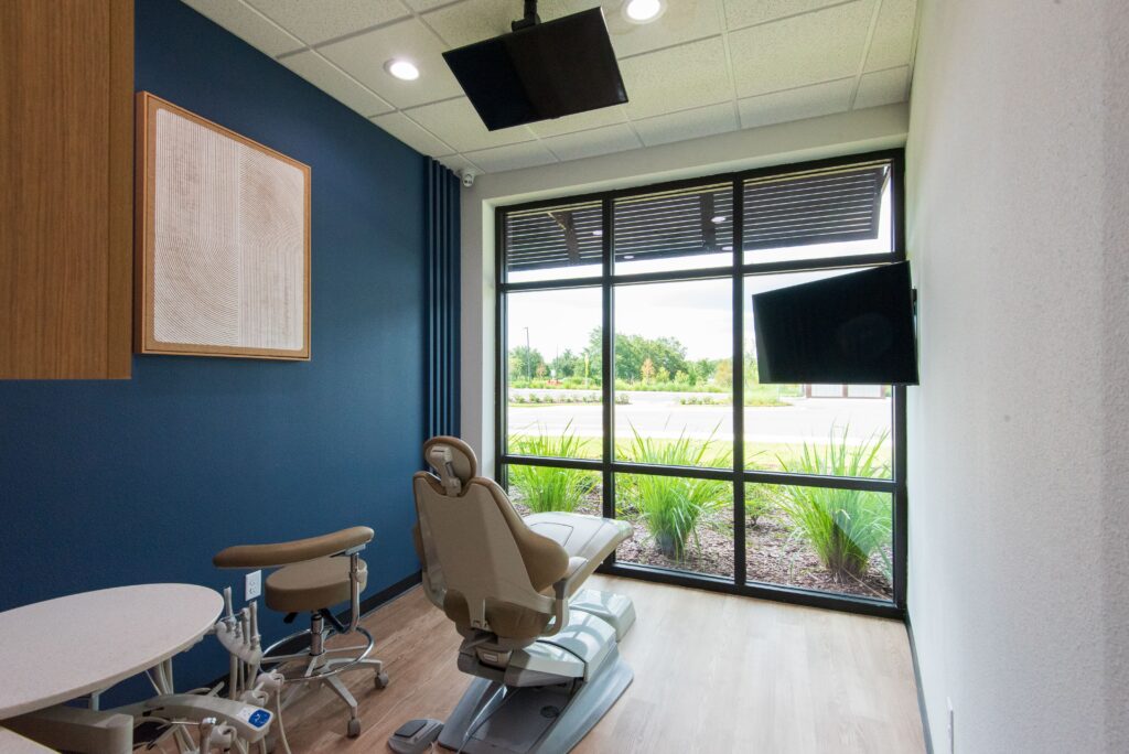 A modern dental exam room with a dental chair, side table, and screen, featuring large windows with a view of greenery outside and blue and white walls.