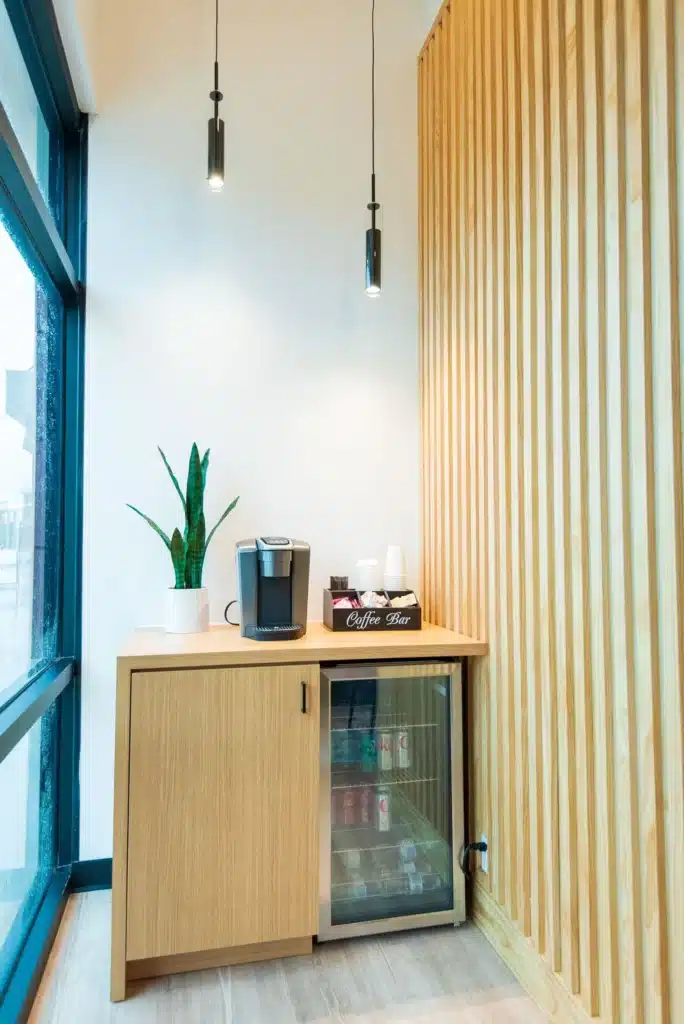 A modern office coffee nook with a Keurig machine, cups, a potted plant, and a mini fridge stocked with drinks, set against wood paneling and lit by two hanging pendant lights.