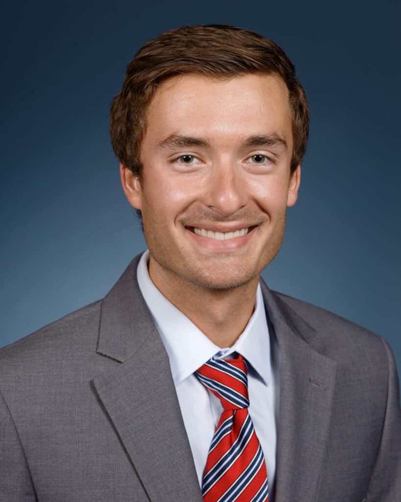 A young man with short brown hair, wearing a light gray suit, white shirt, and red striped tie, smiles at the camera against a plain dark blue background.