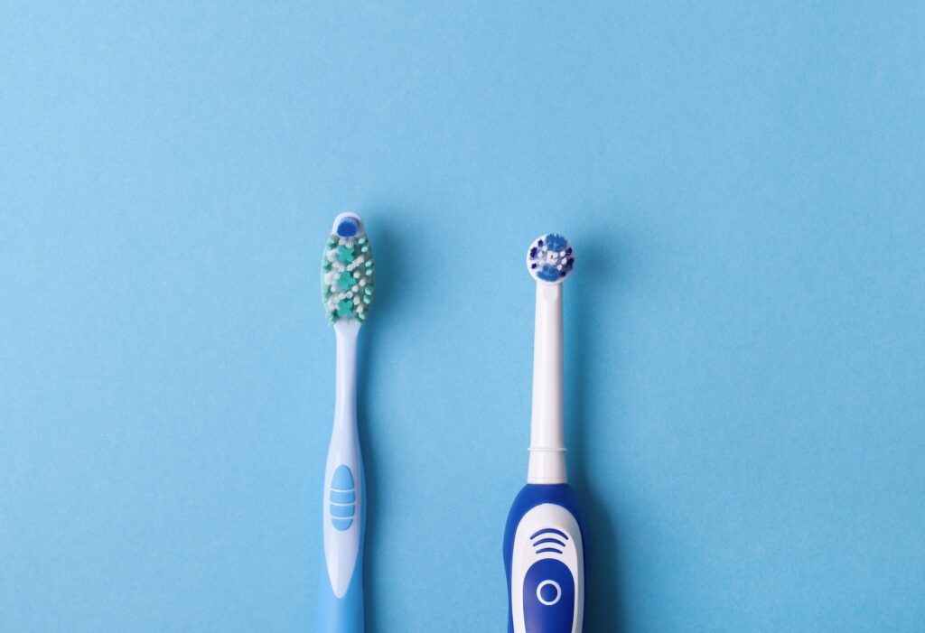 A manual toothbrush with green and white bristles and an electric toothbrush with a round blue and white head are side by side on a light blue background.