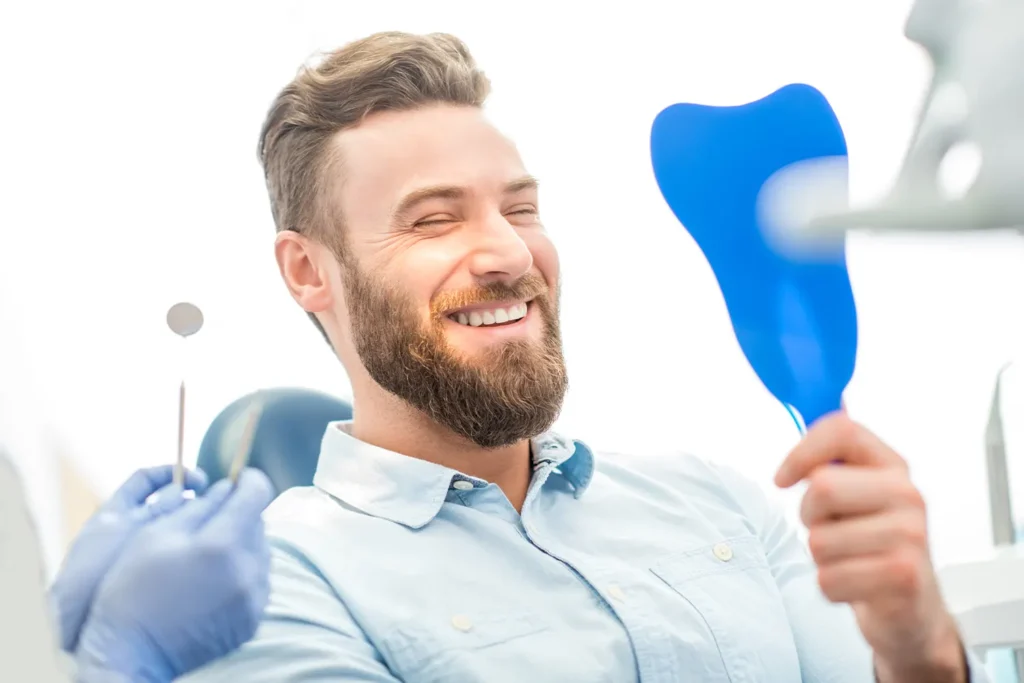 A bearded man smiles while sitting in a dental chair, holding a blue tooth-shaped mirror. A gloved dental professionals hand holds dental tools next to him. The atmosphere appears bright and cheerful.