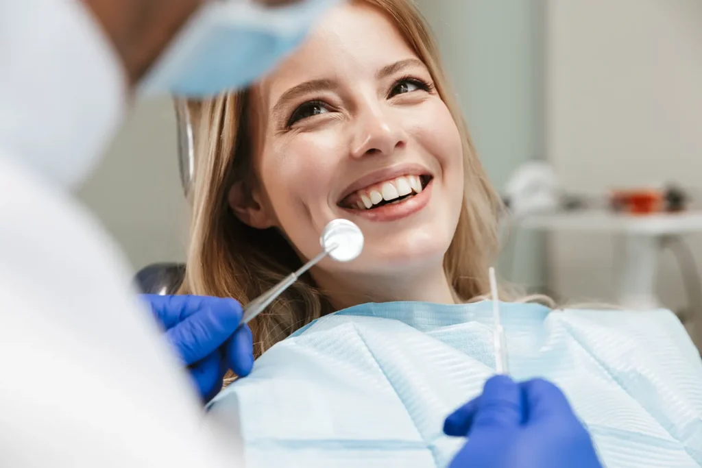 A smiling woman sits in a dental chair while a dentist wearing blue gloves examines her teeth with a dental mirror and tool.