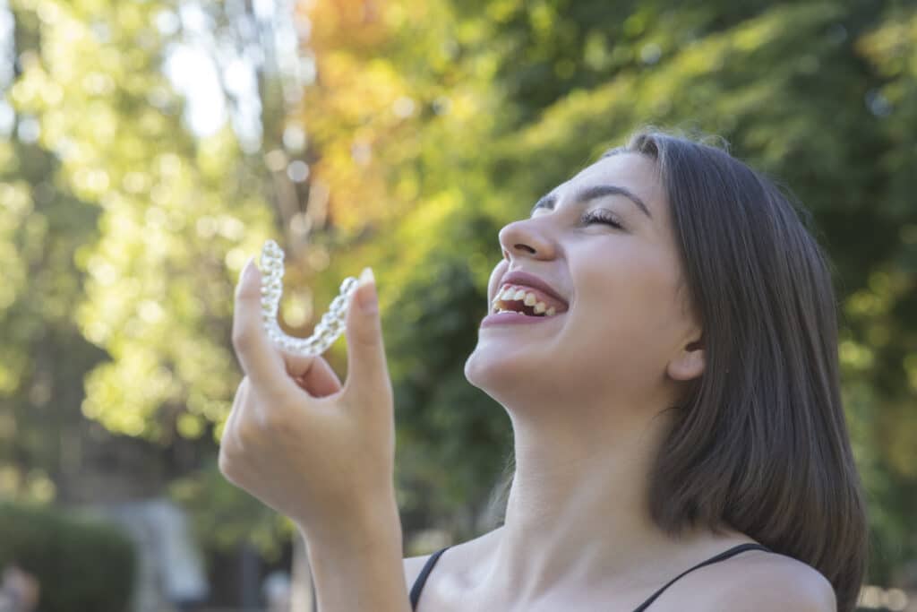 A smiling woman with straight brown hair holds a clear dental aligner outdoors, with blurred green trees and sunlight in the background.