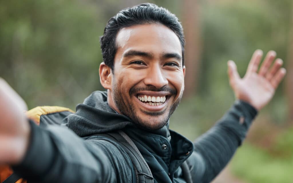 A man outdoors smiles widely at the camera with arms outstretched, wearing a dark jacket and carrying a backpack. The background is blurred with greenery, suggesting a natural setting.
