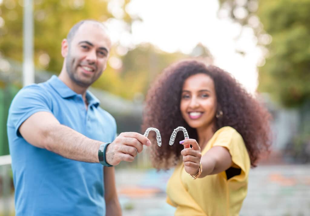 A smiling man and woman stand outdoors, each holding a clear dental aligner towards the camera. The background is blurred with greenery and sunlight.
