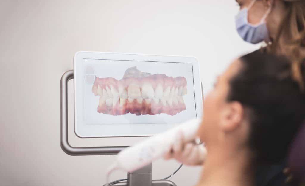 A dentist wearing a mask shows a patient a 3D digital scan of teeth displayed on a monitor during a dental examination. The patient holds a scanning device near their mouth.