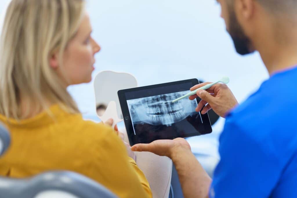A dentist shows a patient a dental X-ray on a tablet, pointing at the image with a toothbrush while explaining. The patient listens attentively.