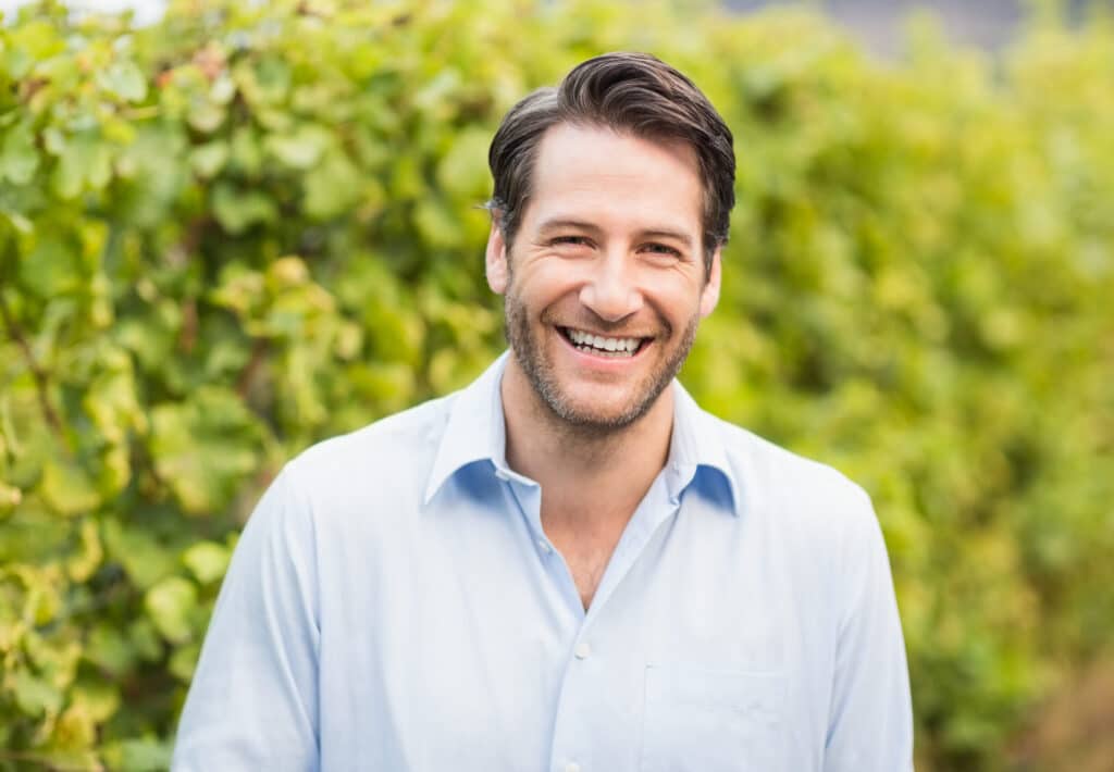 A smiling man with dark hair and a light beard wearing a light blue shirt stands outdoors in front of green leafy vines.