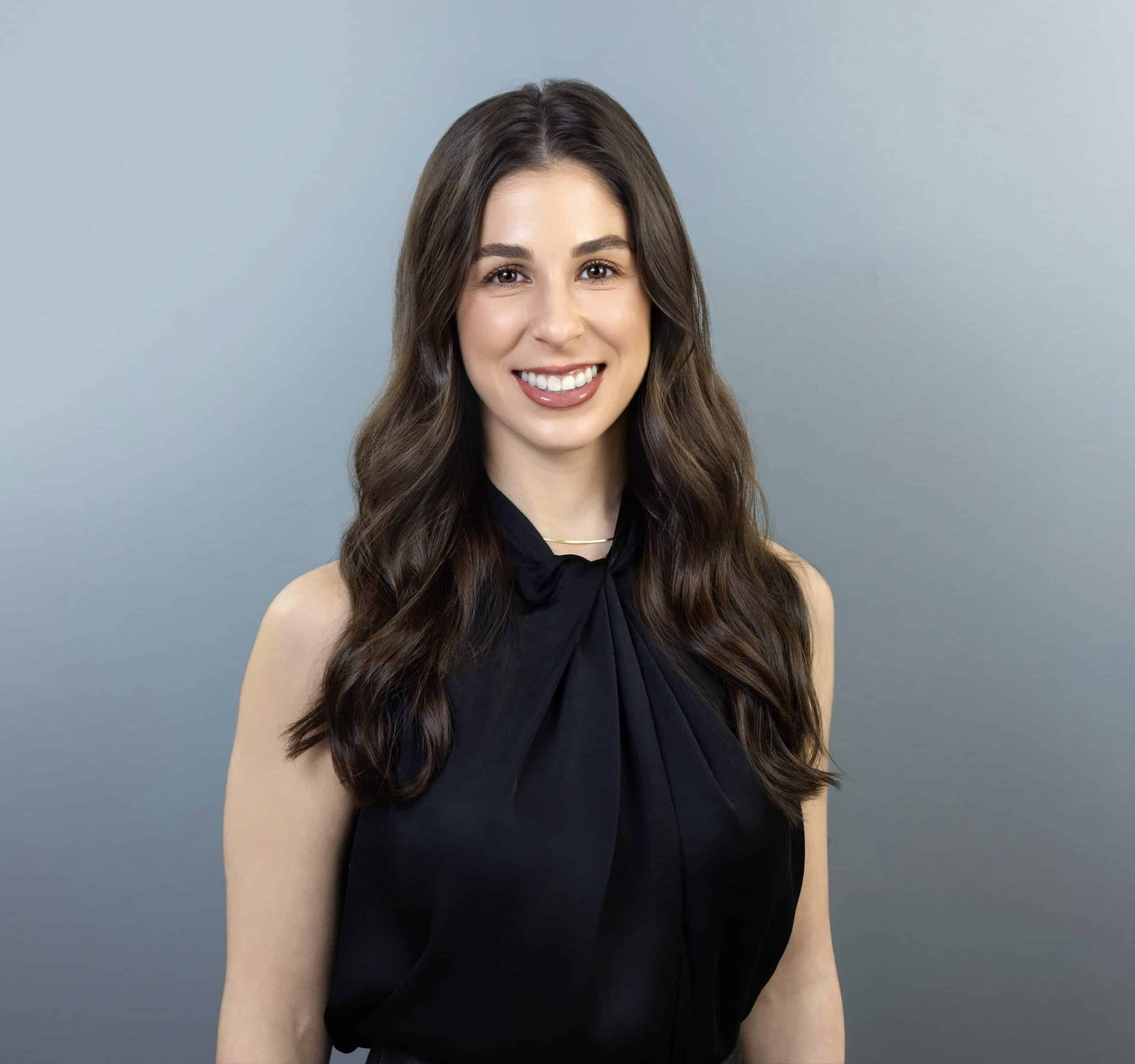 A woman with long, wavy brown hair smiles at the camera. She is wearing a sleeveless black top and standing in front of a plain gray background.